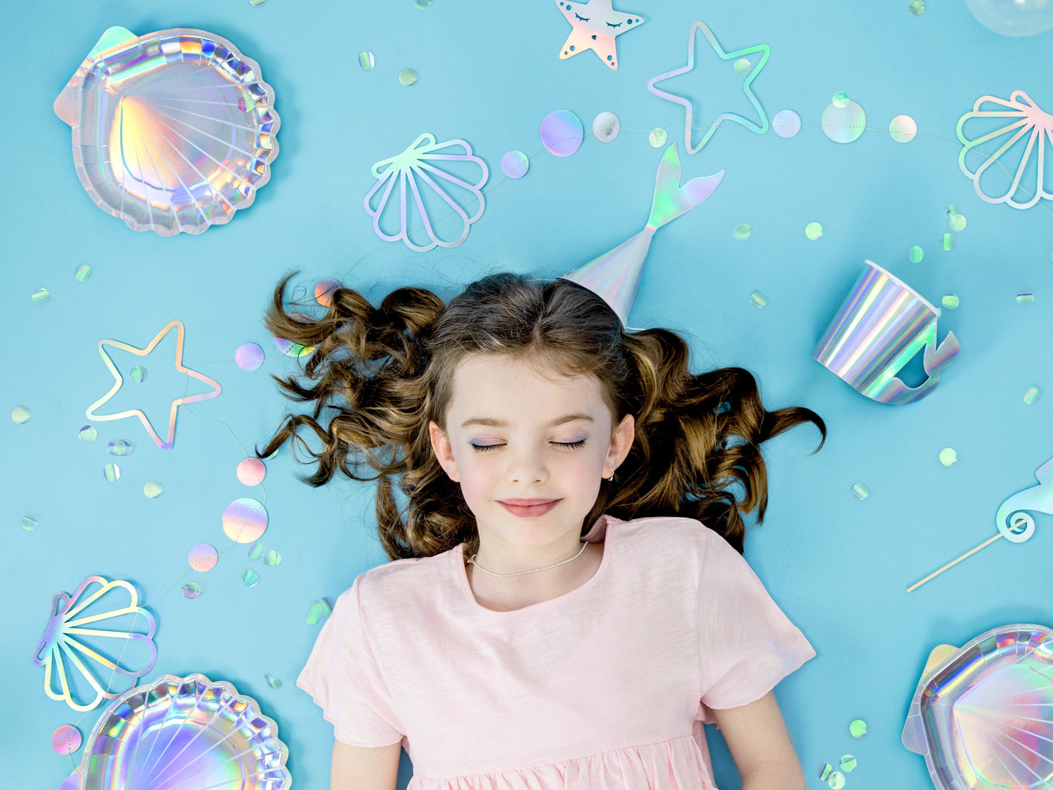 Mermaid birthday party setup with pastel mermaid and seahorse balloons, pink seashell balloon, iridescent decor, jellyfish streamers, 'Happy Birthday' banner and pink table with themed cake