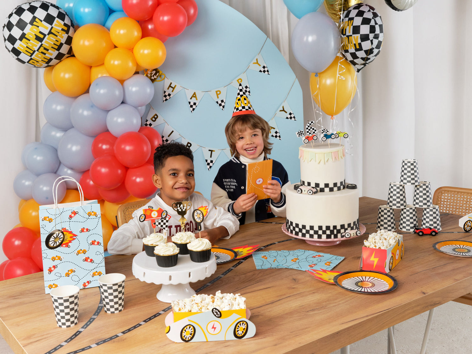 Racing car birthday party setup with colorful balloons, checkered flags, racing car foil balloon, snack boxes, tableware, and cake decorated with race-themed toppers and party hats.