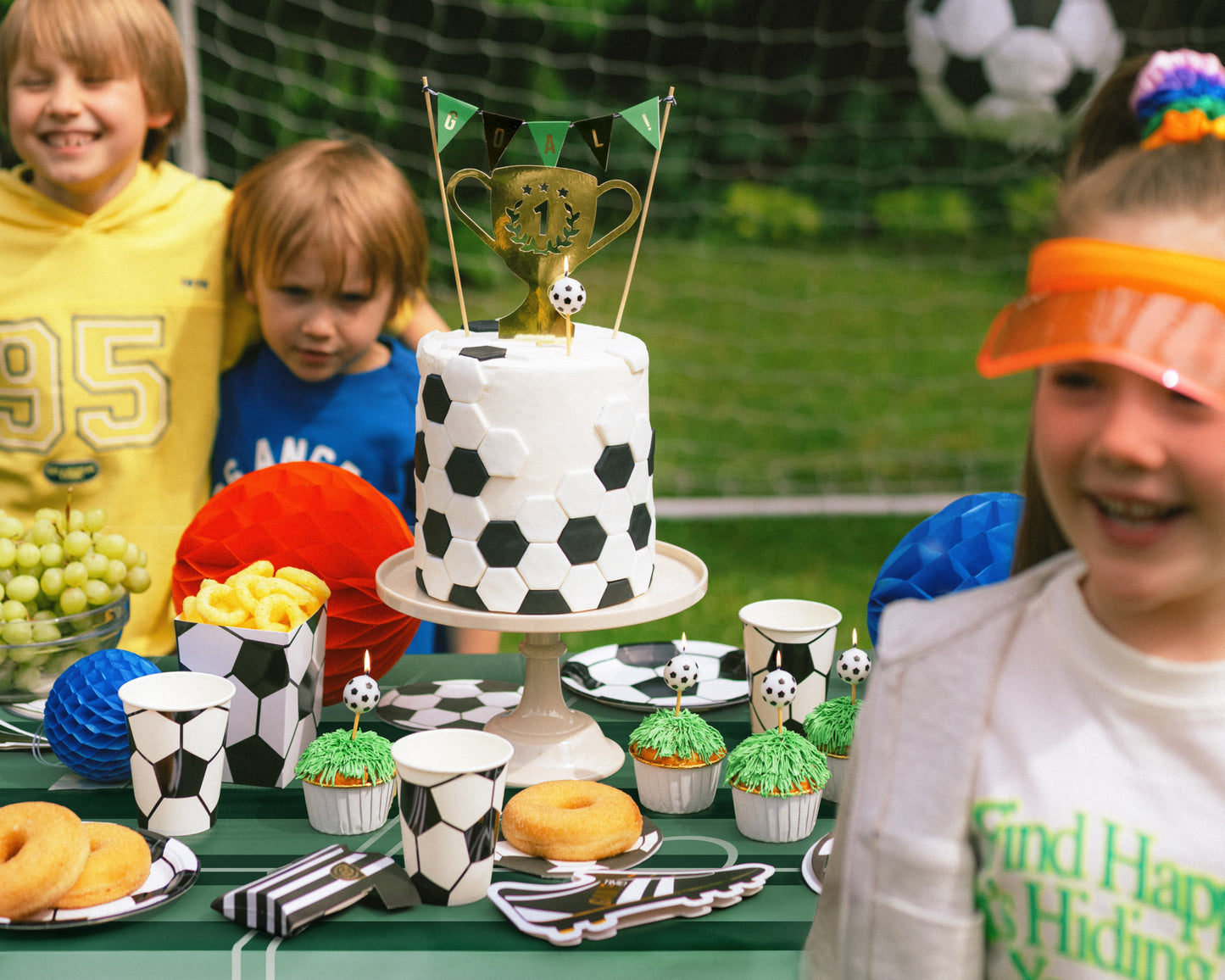 Children around a table with a soccer-themed cake and snacks outdoors.
