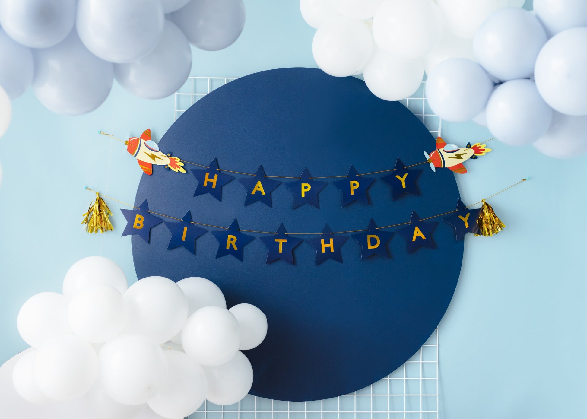 Rocket-themed 'Happy Birthday' banner with navy star letters and gold tassels, surrounded by white and light blue balloon cloud