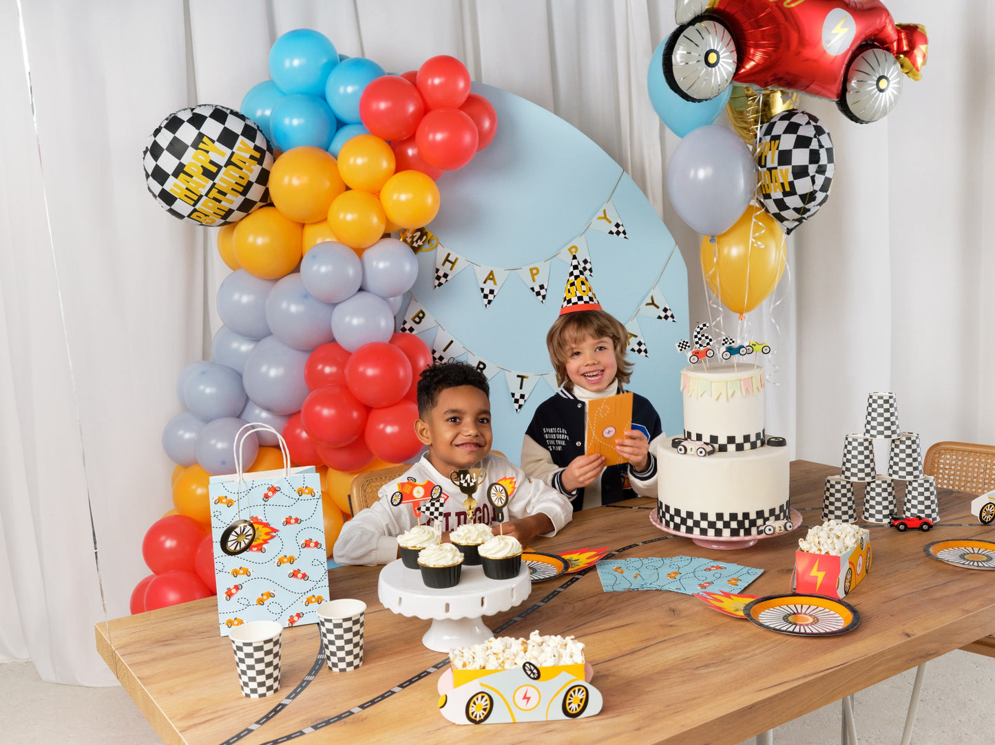 Racing car themed party table with checkered paper cups, wheel-shaped plates, popcorn snack boxes shaped like race cars, muffins, and colorful flame decorations.