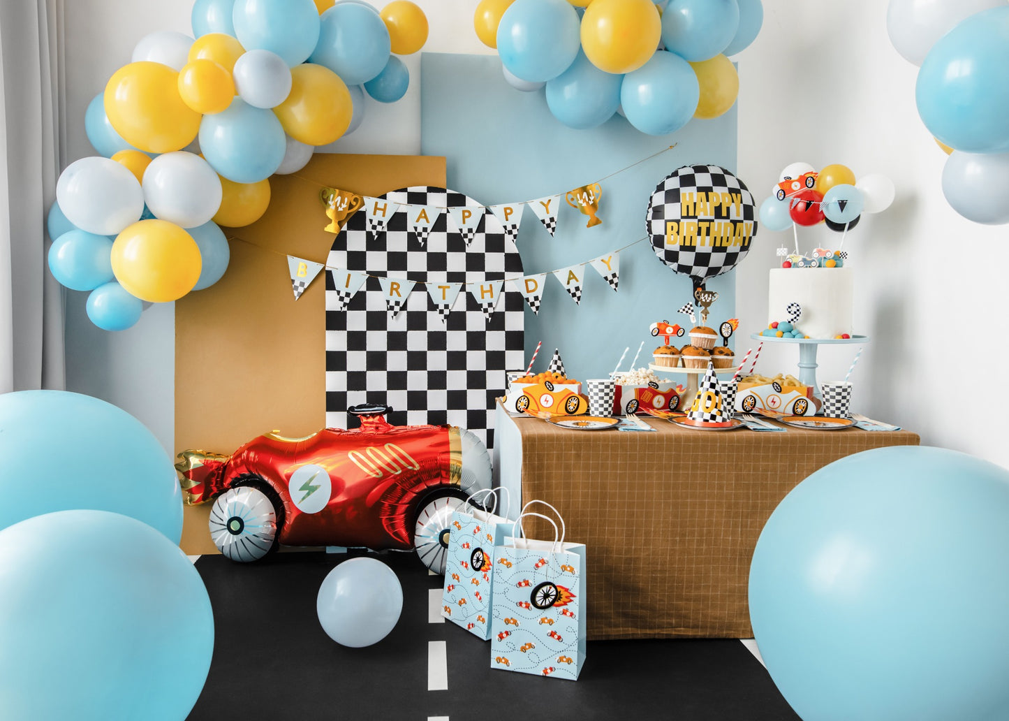Racing car birthday party setup with colorful balloons, checkered flags, racing car foil balloon, snack boxes, tableware, and cake decorated with race-themed toppers and party hats.