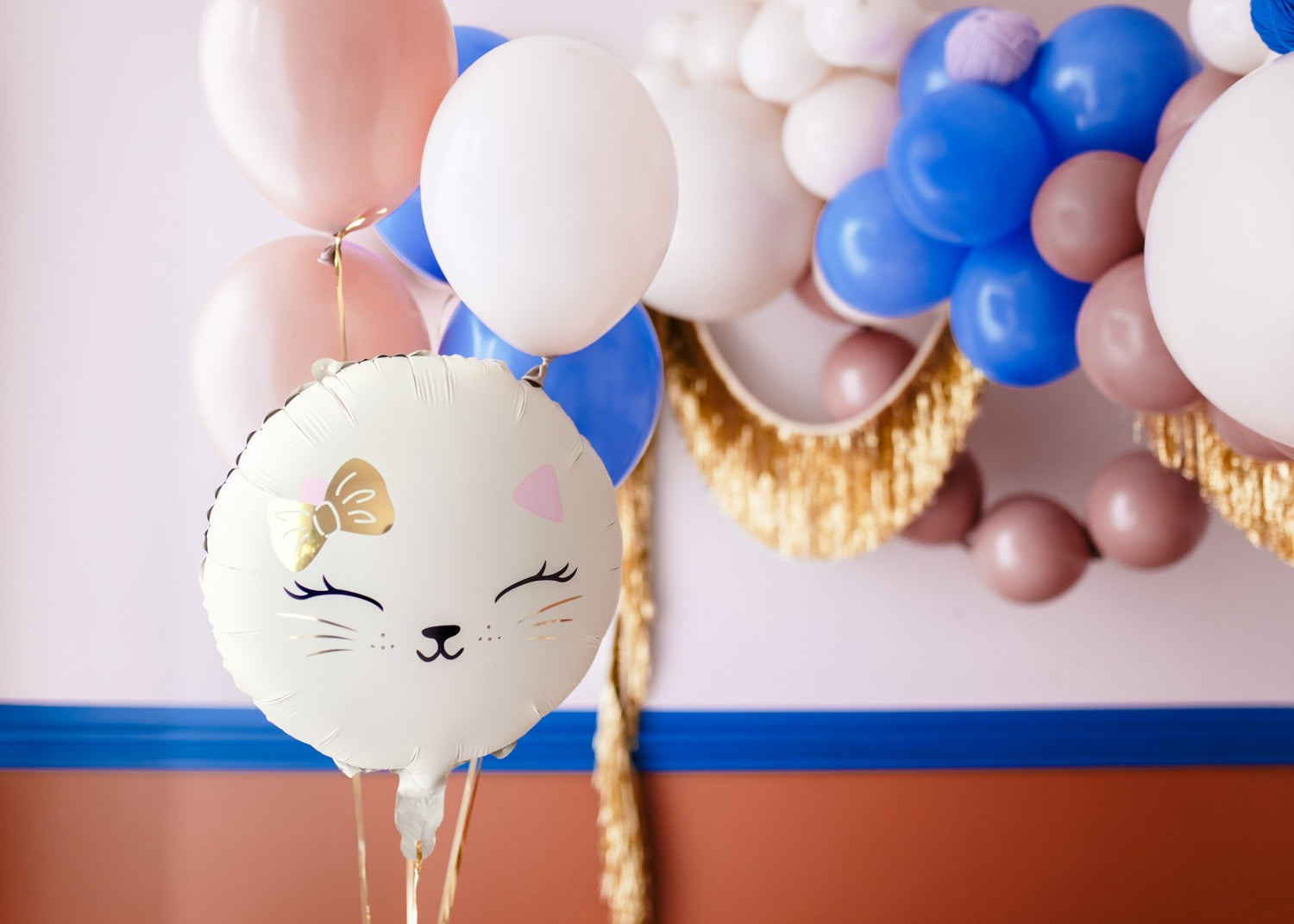 Cat-themed birthday party decorations with a "Happy Birthday" banner, pastel and metallic balloons, gold garland, cat face gift bags, snack boxes, and pink cake on a festive table, perfect for a kids' kitty party.