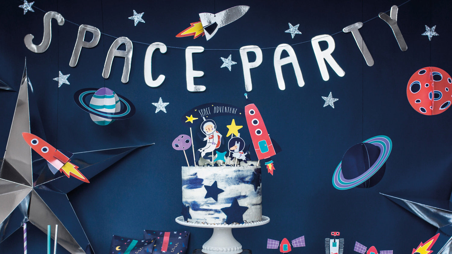 Space-themed birthday dessert table with rocket and planet decorations, SPACE PARTY banner, galaxy cake toppers, silver star balloons, space napkins, and metallic party cups