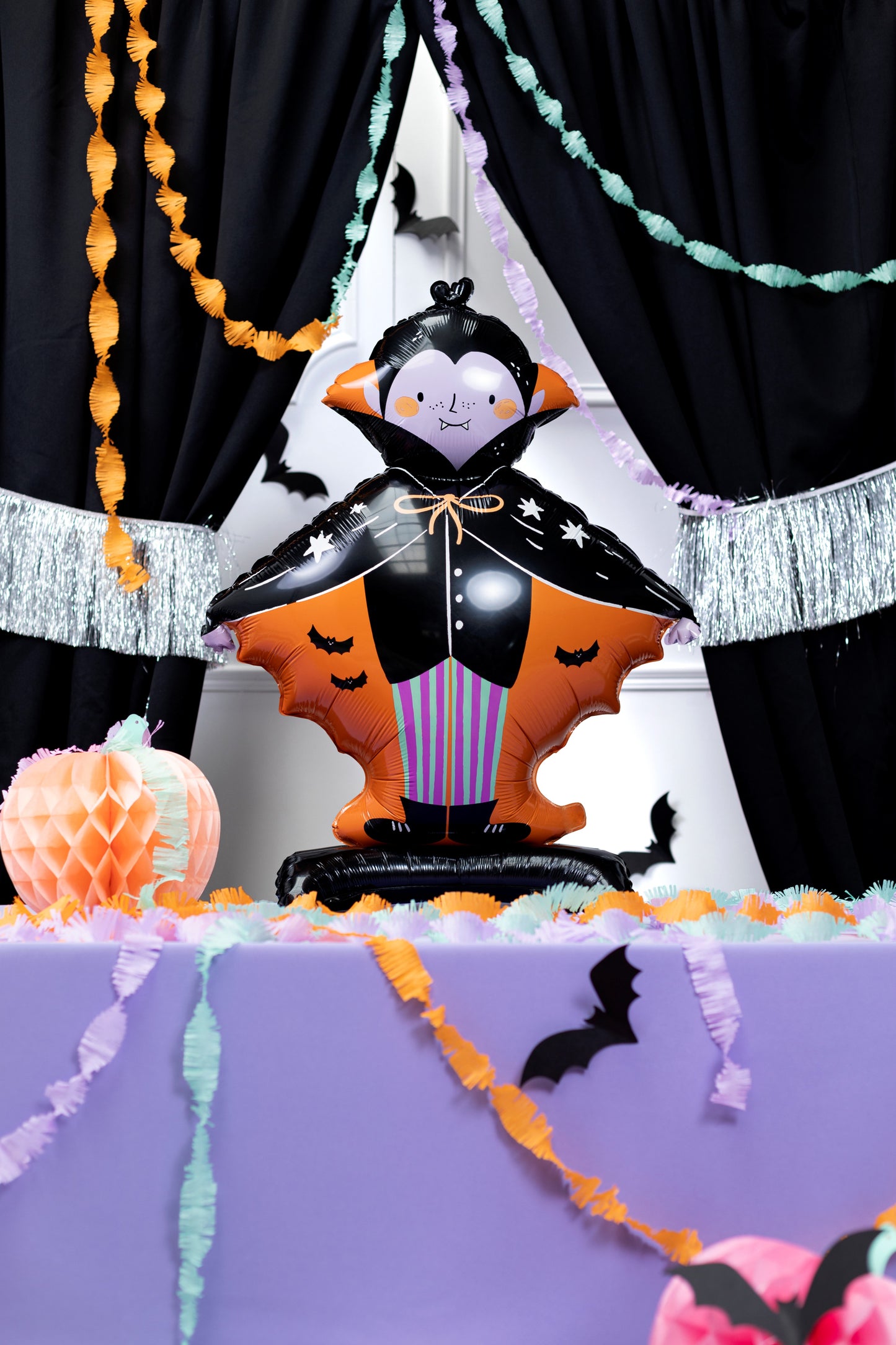 Halloween-themed decoration with a vampire balloon, pumpkins, and black bats on a purple tablecloth.