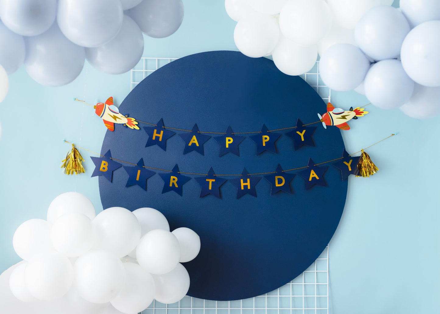 Rocket-themed 'Happy Birthday' banner with navy star letters and gold tassels, surrounded by white and light blue balloon cloud