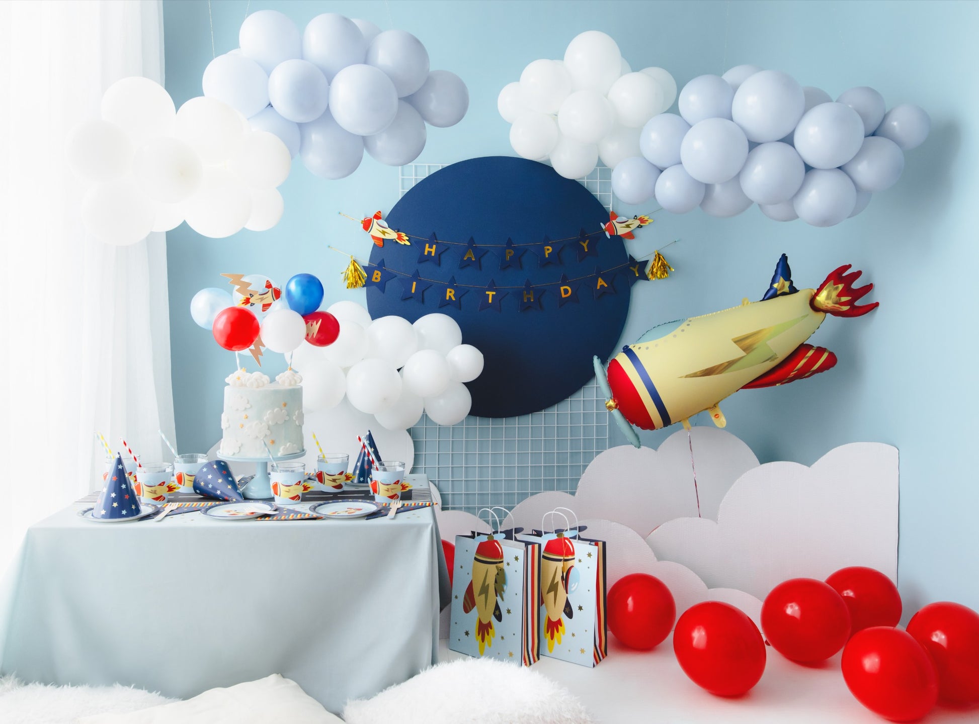 Complete rocket-themed birthday setup with cake, red balloons, foil rocket balloon, and decorated table surrounded by clouds