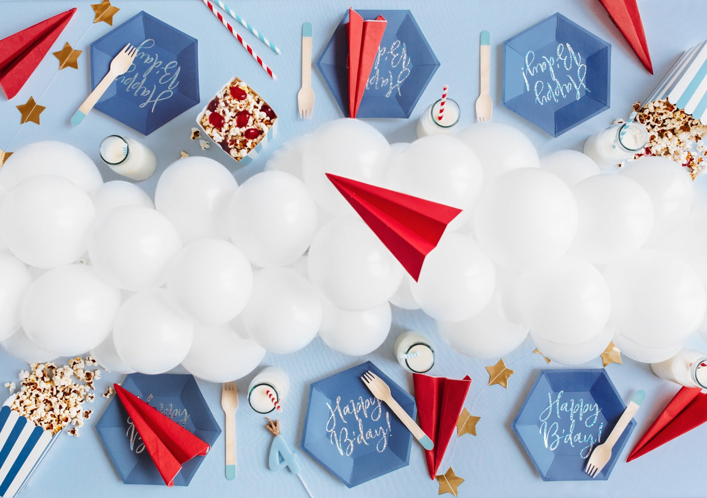 Aviation-themed birthday party table setup with blue hexagon 'Happy Birthday' plates, red paper planes, white balloon cloud centerpiece, and striped popcorn boxes – airplane party decoration for kids