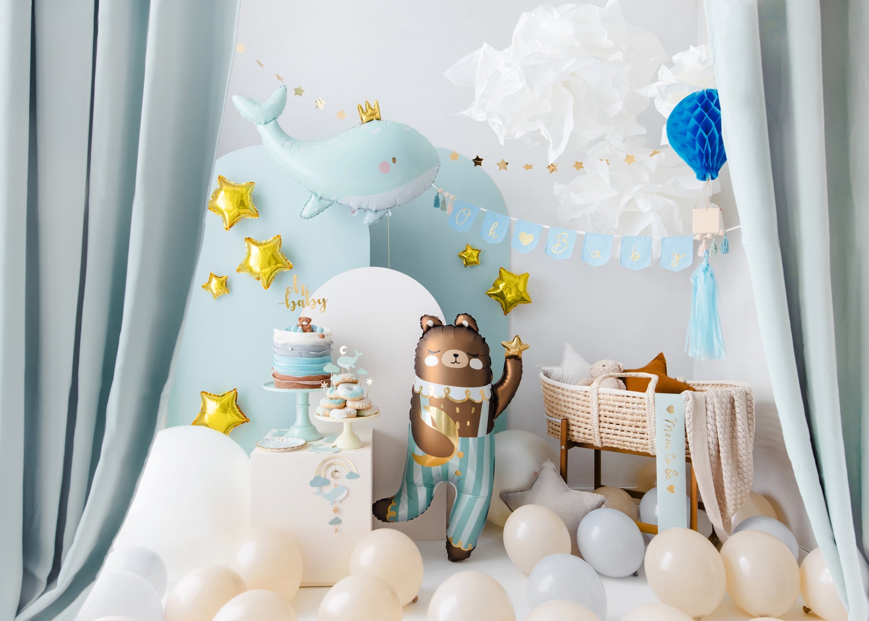 Baby boy welcome party setup in soft blue tones with whale and teddy bear foil balloons, Oh Baby banner, star decorations, cake table, and bassinet – perfect for a newborn baby shower.