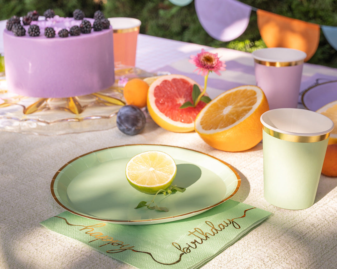 Table setting with a cake, fruit, and 'Happy Birthday' napkin on a light surface.